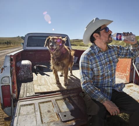 man drinking ozarka water in the back of a pickup truck with his dog